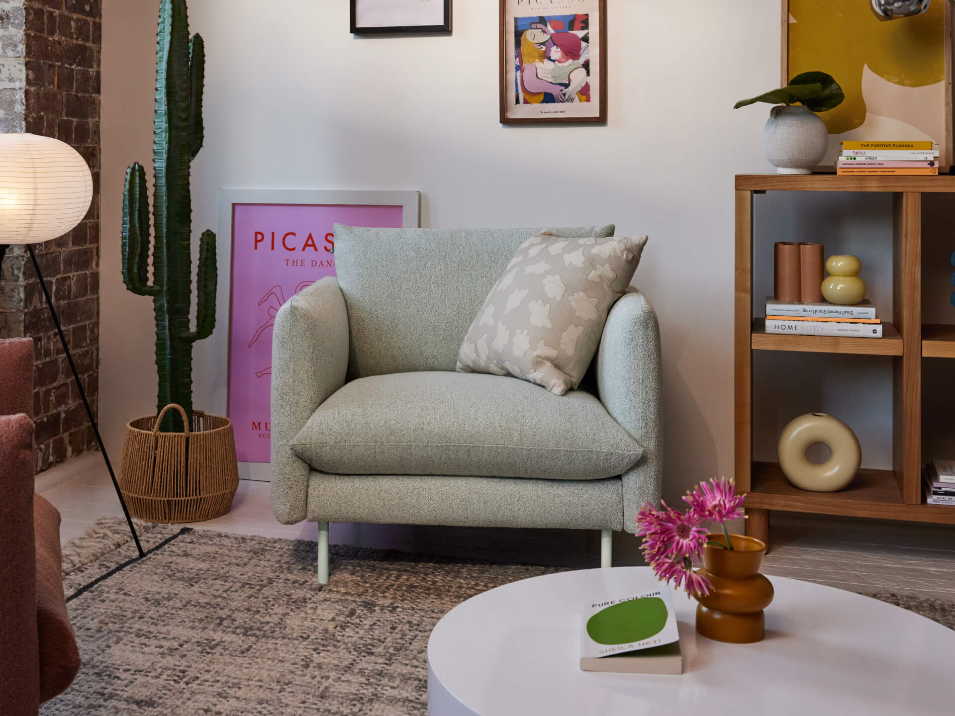 Light grey armchair with a patterned cushion in a stylish living room. Features include a cactus, framed prints, and a wooden shelf with decor items.