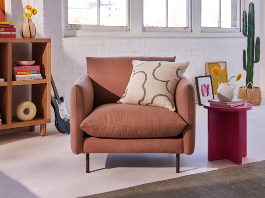 A cosy brown armchair with a patterned cushion, set on a light floor beside a modern wooden shelf and a cactus near a magenta side table.