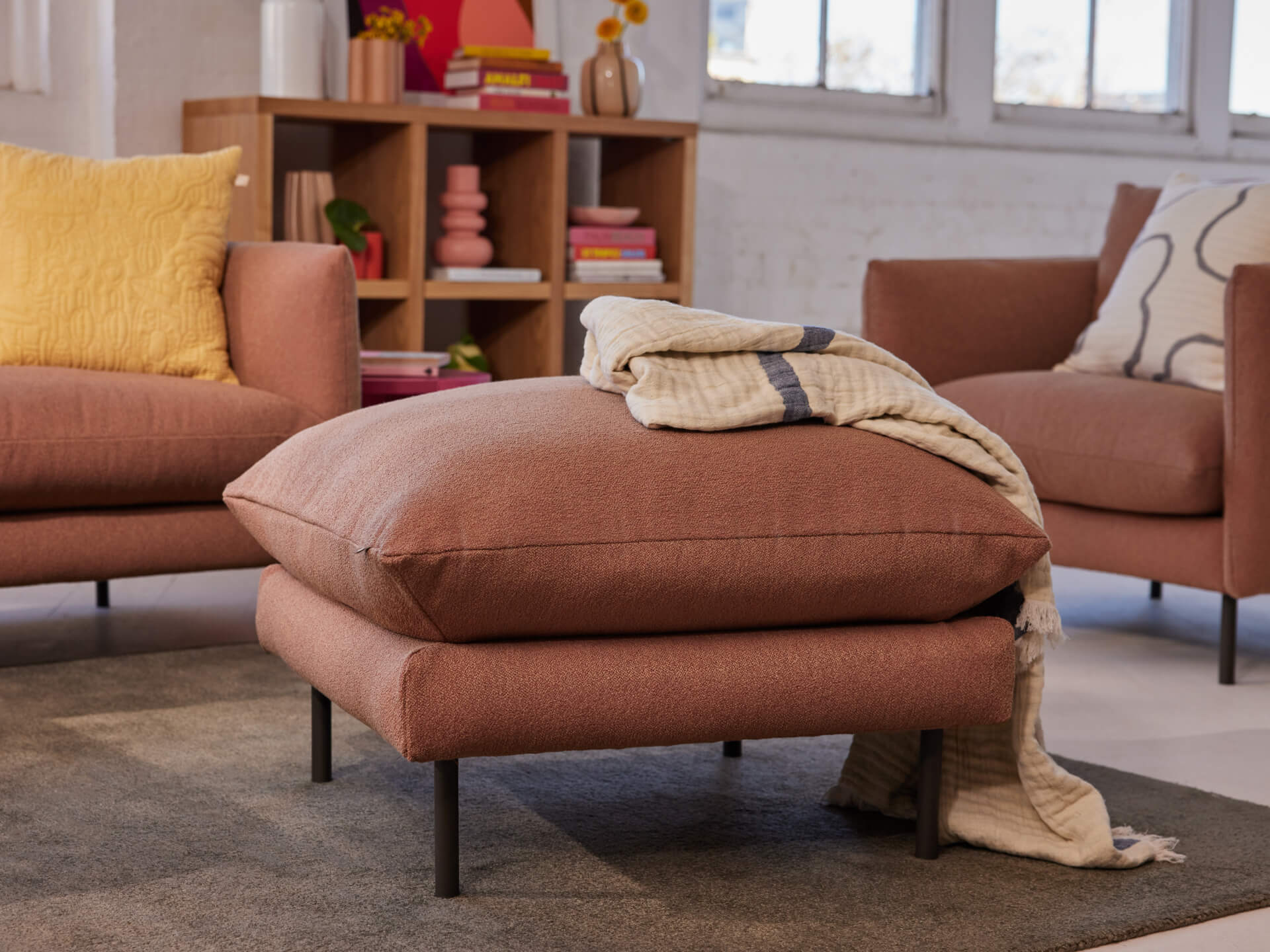 Rust-coloured ottoman with black legs, a beige throw draped over it. Two matching armchairs, yellow cushion, and a shelf in the background.