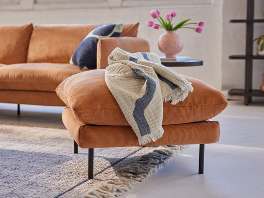 A brown sofa with a matching ottoman, draped with a cream and navy throw. Nearby, a pink vase with tulips sits on a black side table.
