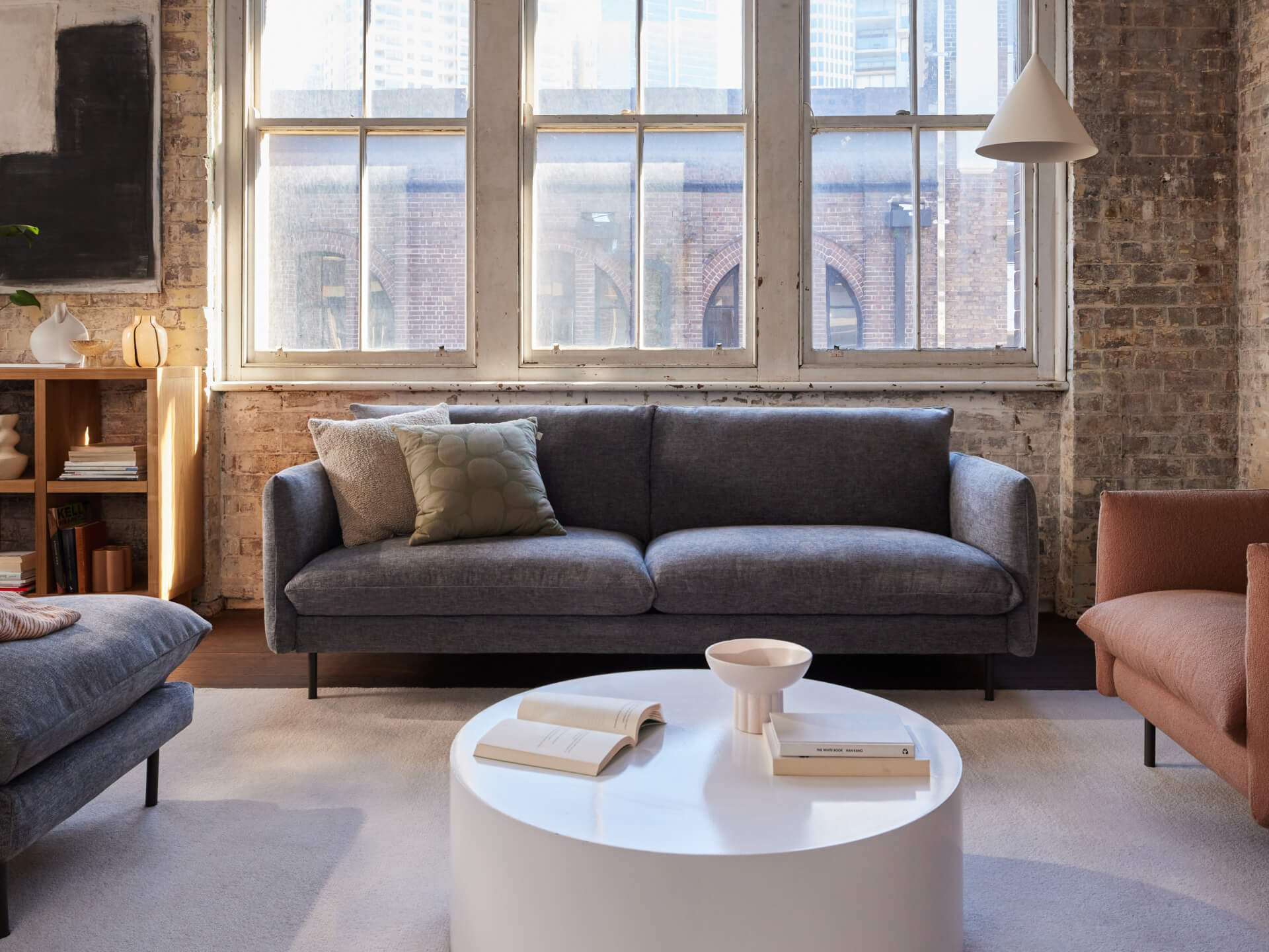 Living room with a dark grey sofa, brown armchair, round white coffee table, and books, set against exposed brick walls and large windows.