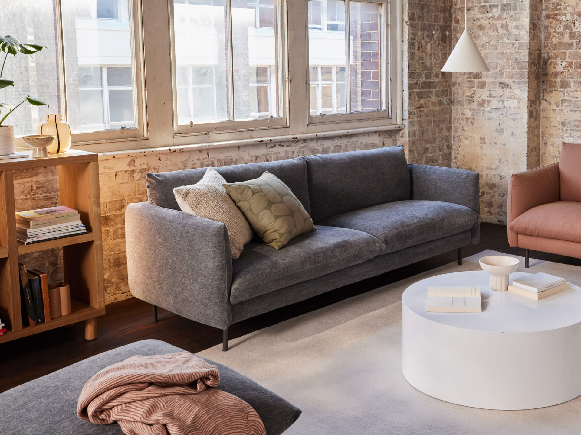 Loft-style living room with a dark grey sofa, beige cushions, white round coffee table, and a tan armchair. Exposed brick walls and tall windows.