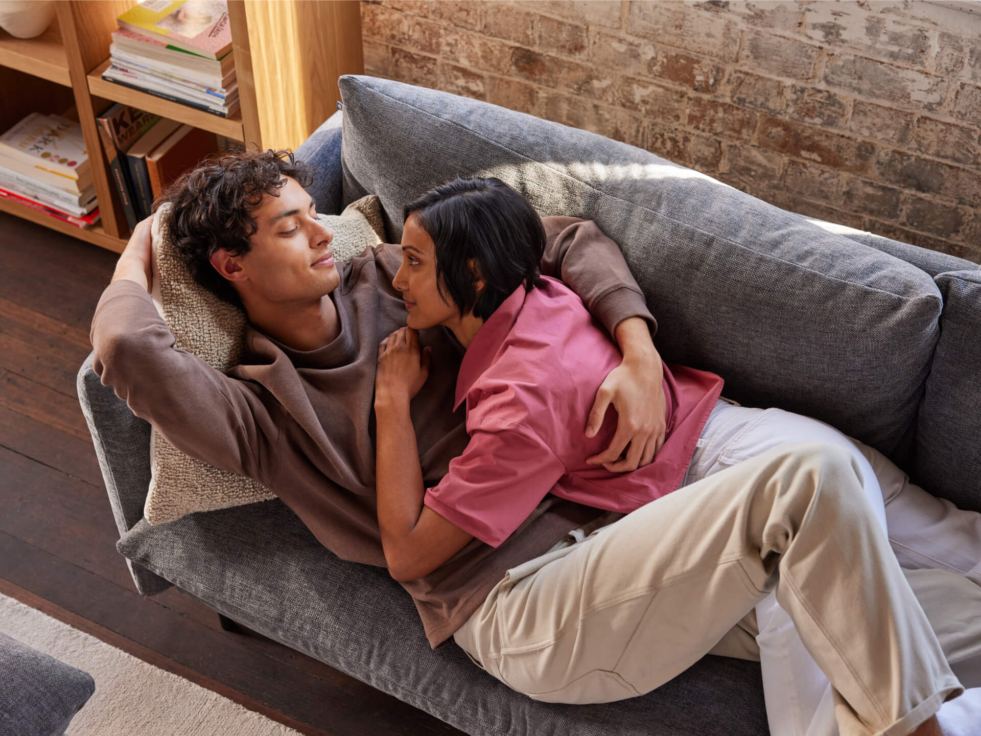 Couple lounging on a grey sofa in a cosy living room with wooden shelves and a textured brick wall.