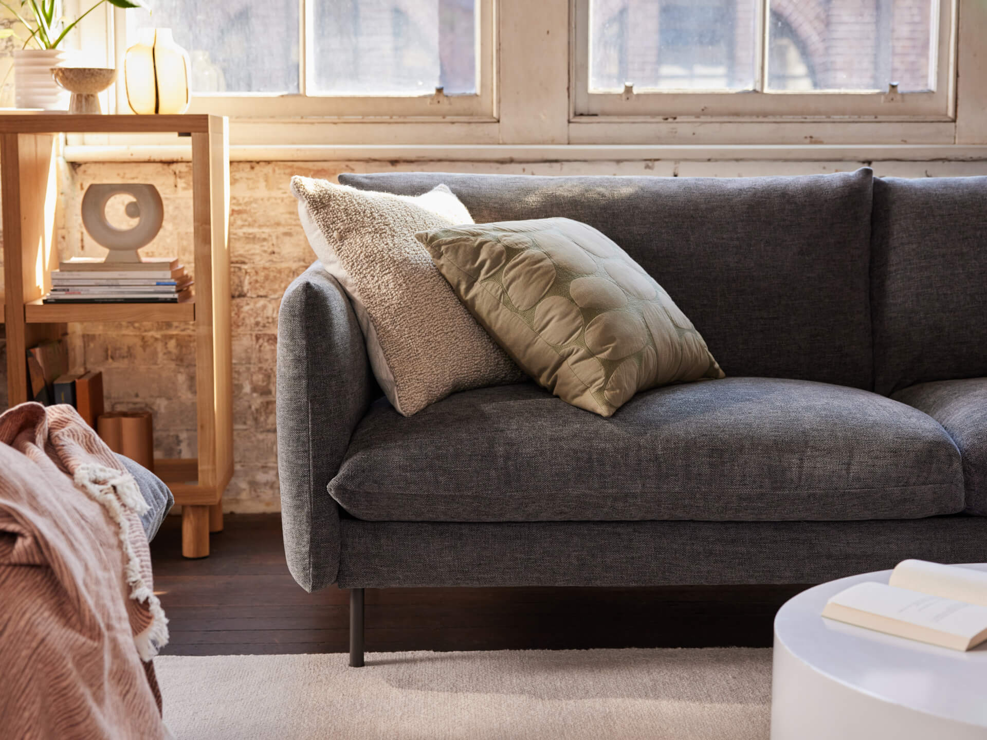 Modern living room with a dark grey sofa, cream and green cushions, wooden bookshelf, and round white coffee table. Warm, natural light from windows.