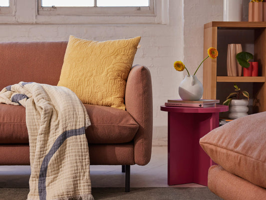 Rust sofa with yellow cushion, draped beige throw, and pink side table with vase. Shelving and pink floor cushion in background. Cozy living space.