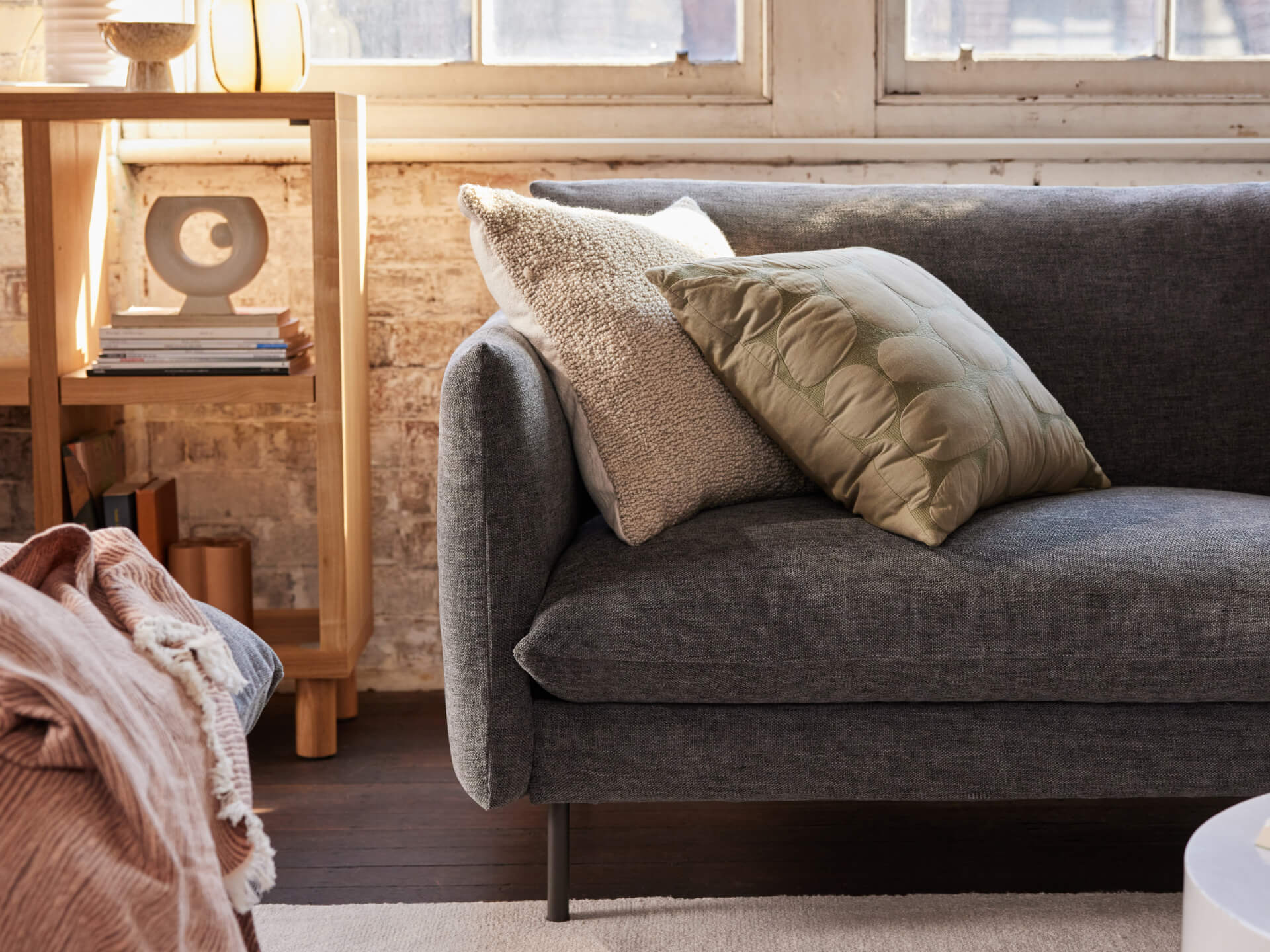 Grey fabric sofa with green and beige cushions, set against a rustic brick wall and wooden shelf. Warm, cozy living room ambience.