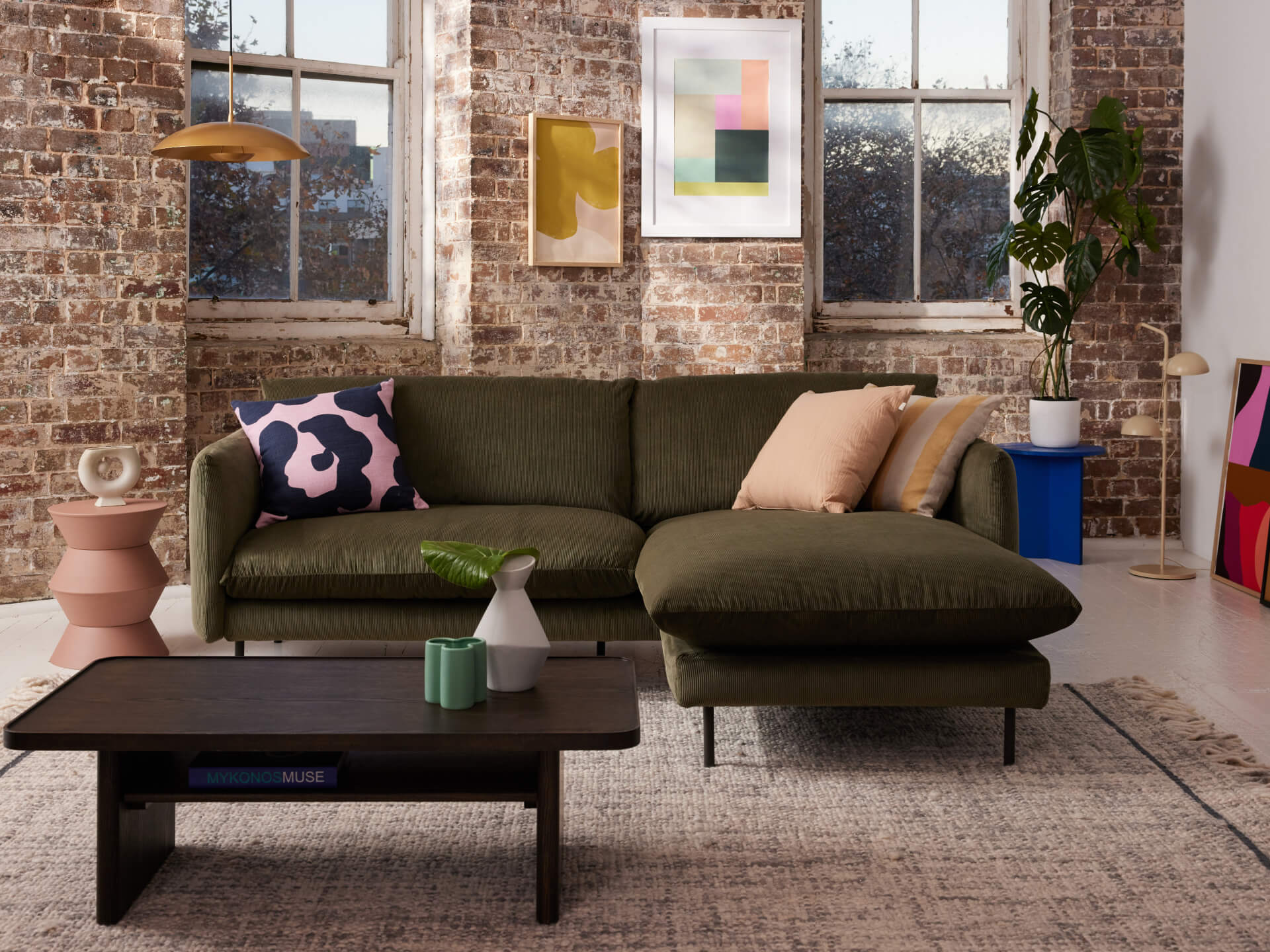 Living room with dark green sectional sofa, multicoloured cushions, black coffee table, indoor plant, brick wall, and abstract art.