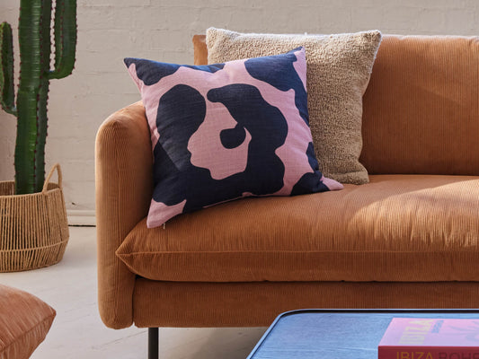 Rust-coloured sofa with a pink and black abstract cushion and a tan textured cushion. Cactus plant on left; coffee table with book in foreground.