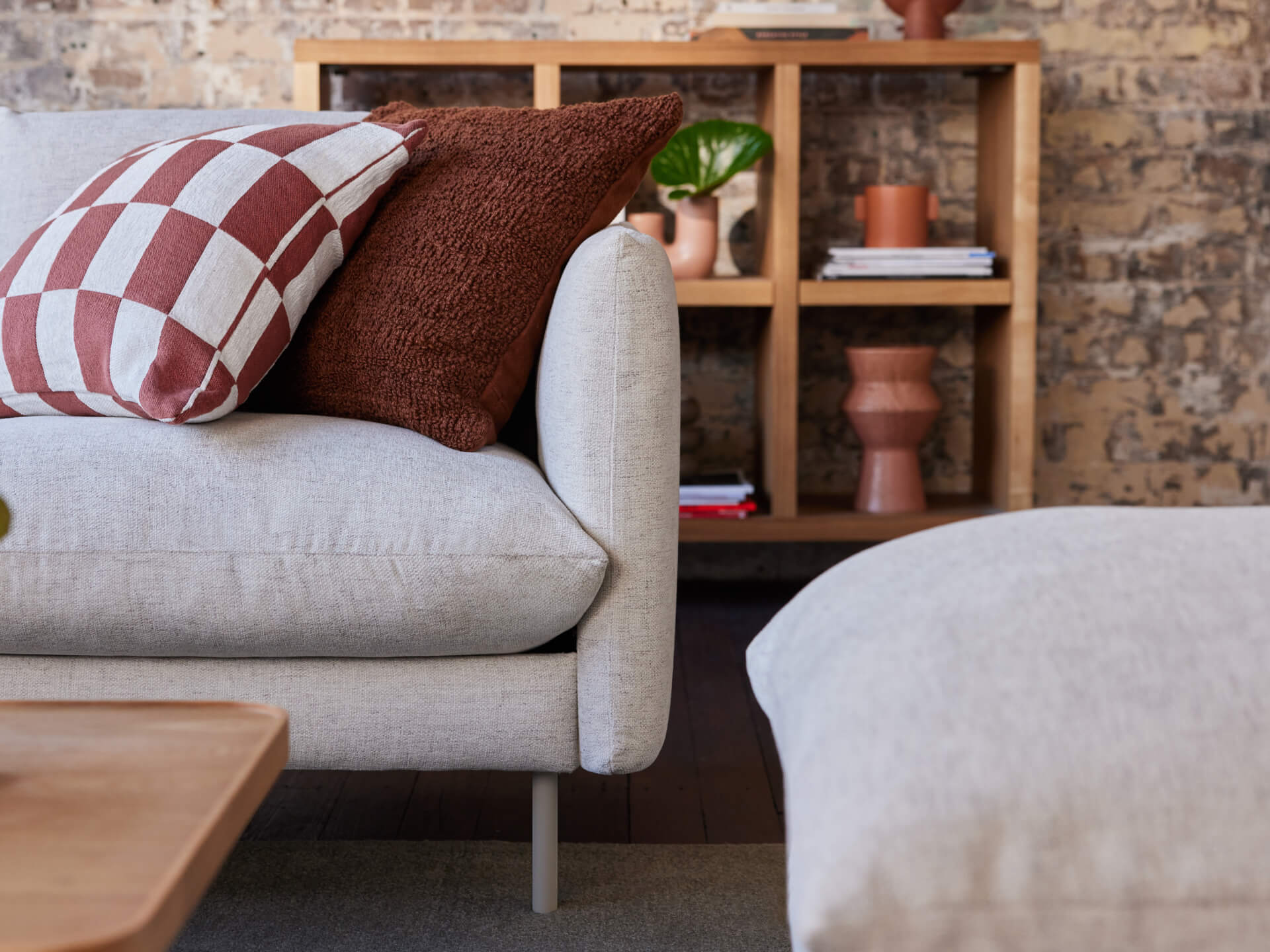 Beige sofa with brown and checkered cushions, in front of a wooden shelf with plants and books, against a brick wall backdrop.