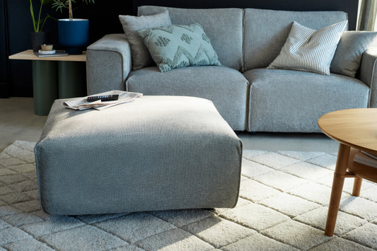 Modern living room with a grey sofa, matching ottoman, light wood coffee table, and a textured rug. Cushions and a plant decorate the space.