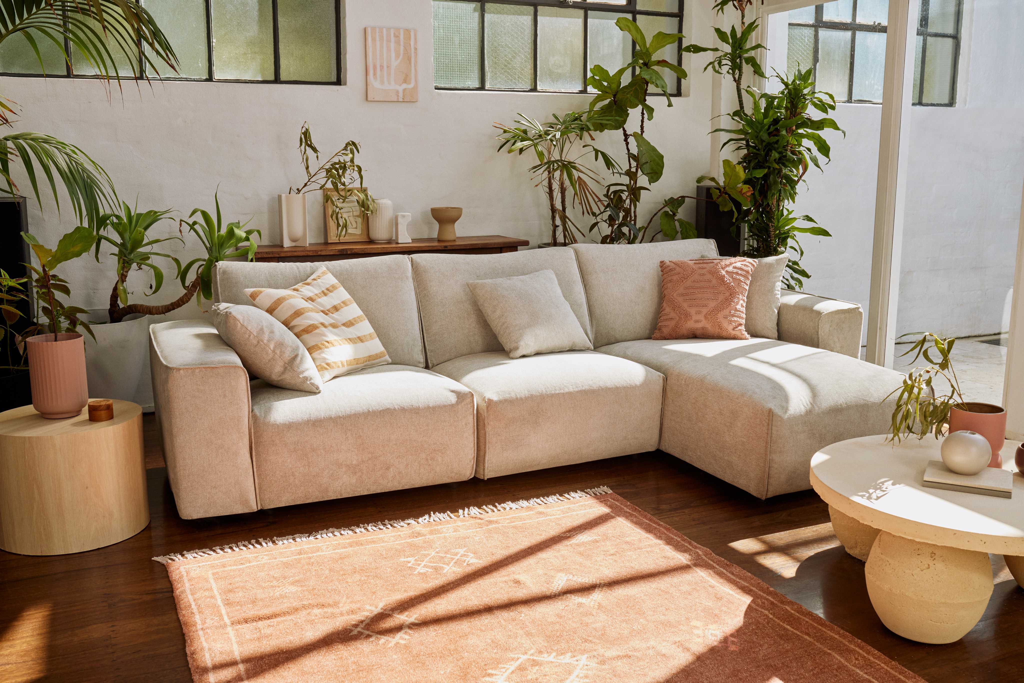 Modern living room with an L-shaped light beige sofa, assorted cushions, wooden coffee table, indoor plants, and large windows. Warm, cosy ambiance.