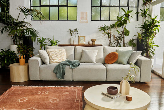 Light grey sofa with cushions in a plant-filled living room, featuring wooden tables and large windows. Boho rug and decor accents complete the scene.