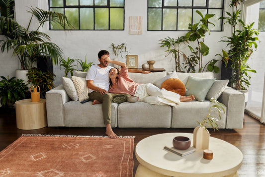 Couple relaxes on light grey sofa surrounded by indoor plants, with coffee table and rust-coloured rug in bright living room.