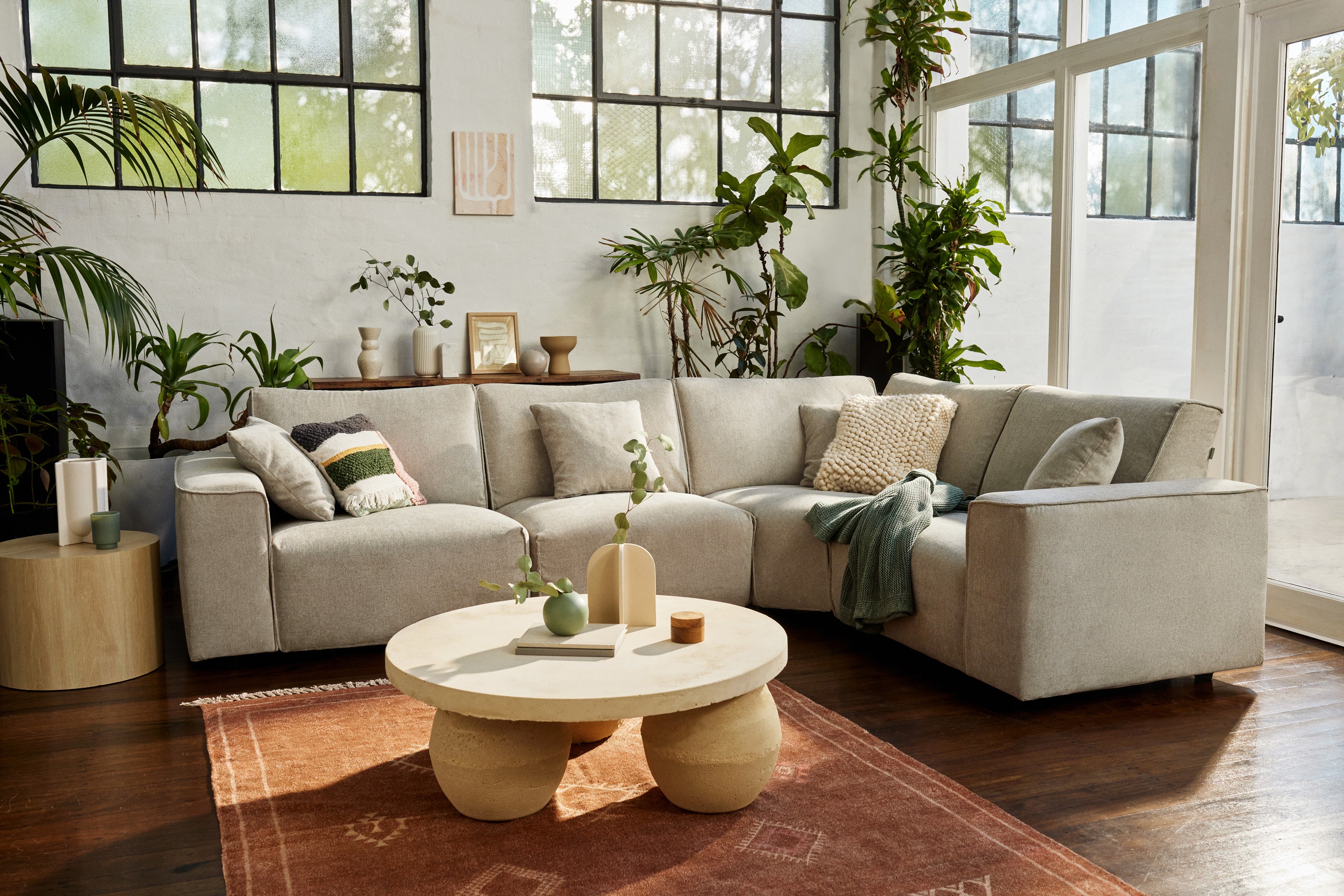 Modern living room with a light grey L-shaped sofa, round wooden coffee table, and indoor plants. Large windows and a terracotta rug enhance natural light.