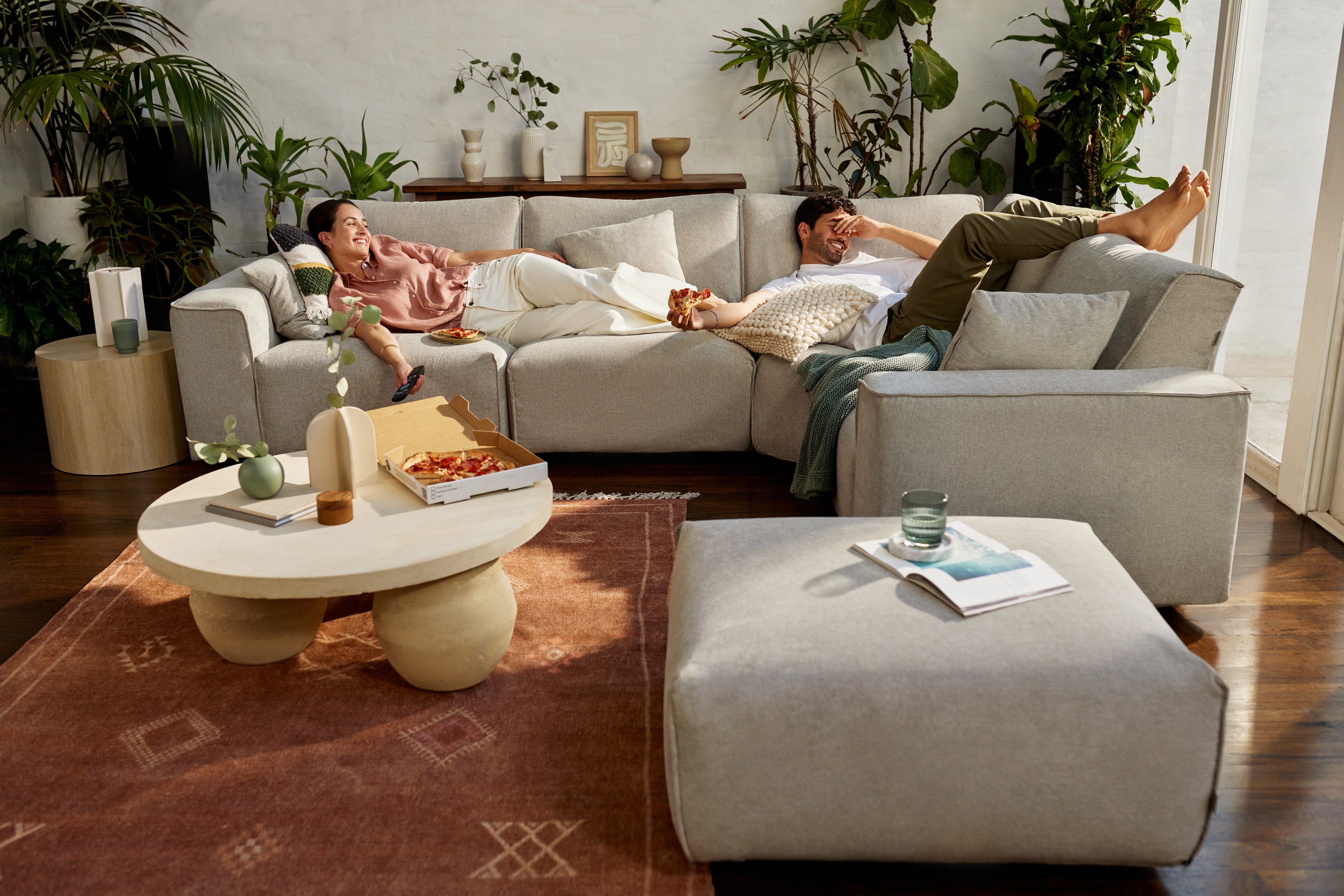 A grey modular sofa set with scattered cushions in a living room. A light brown rug, round coffee table, plants, and two people relaxing.