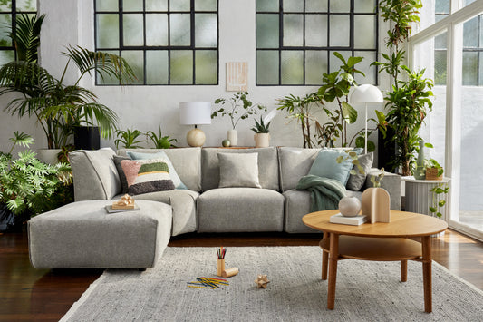 Modern living room with a light grey sectional sofa, wooden coffee table, and lush green plants. Large industrial-style windows and a grey rug.