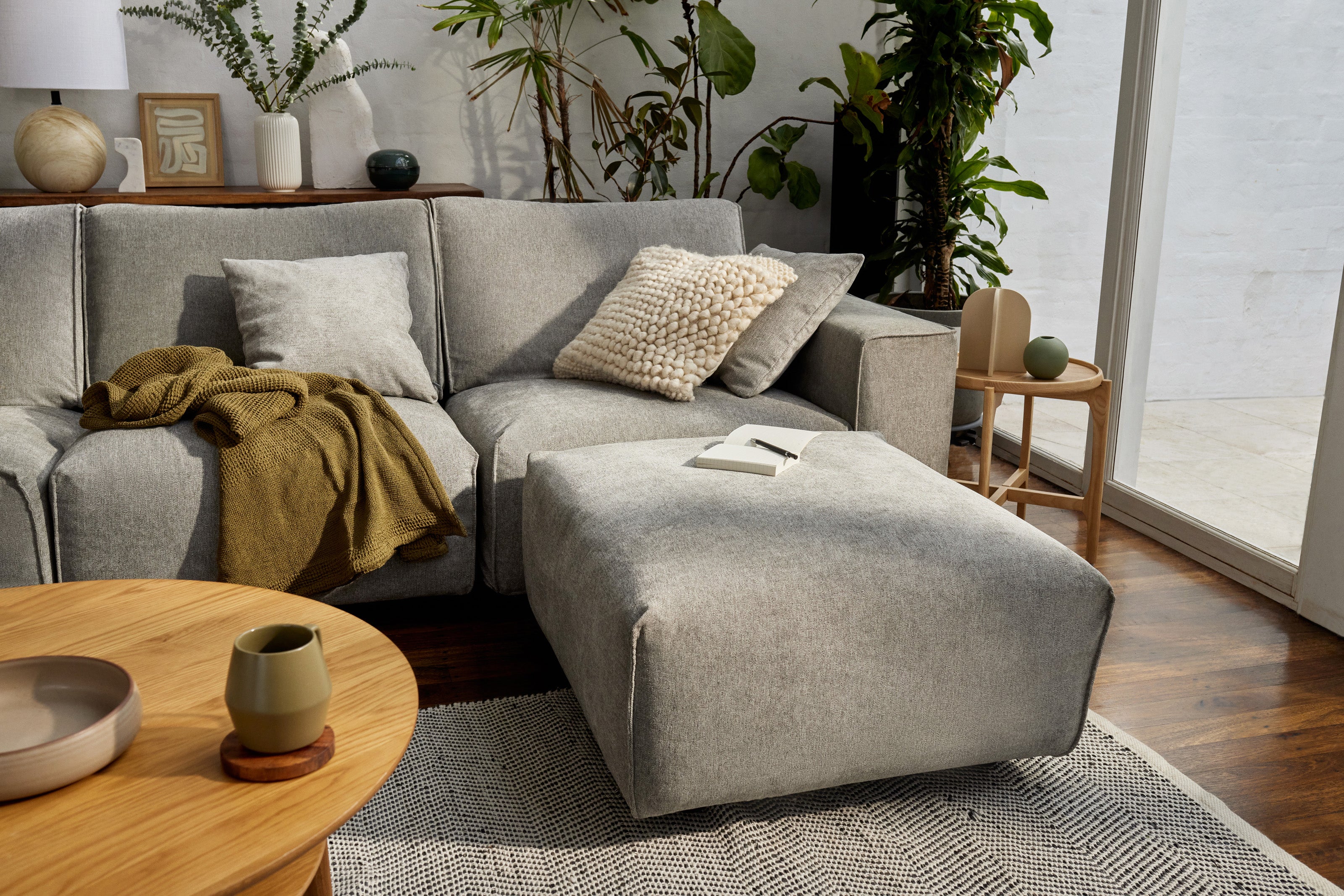 Living room with light grey sectional sofa, textured cushions, olive throw, large ottoman, wooden coffee table, and plants by glass doors.