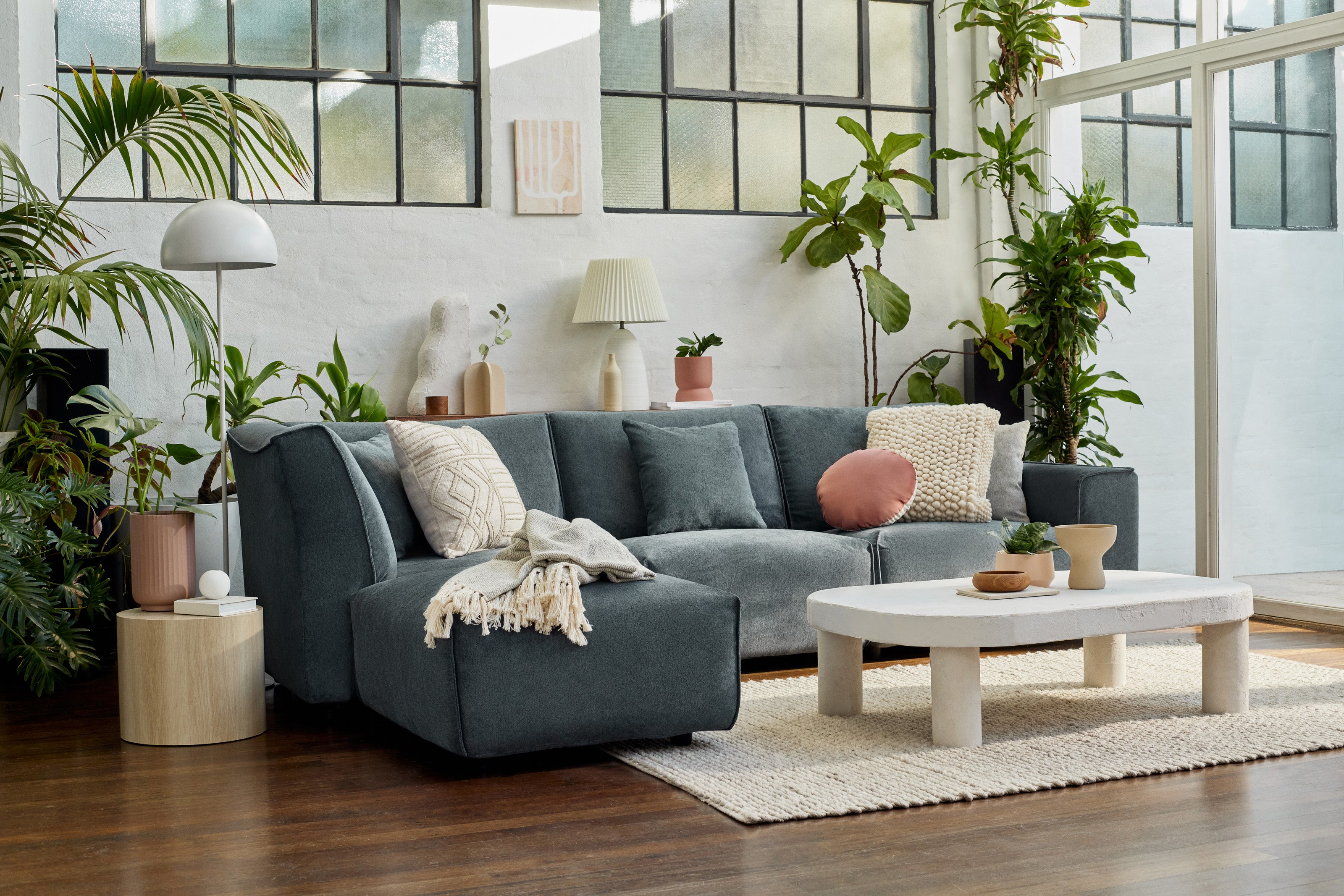 A cosy living room with a dark grey sectional couch, assorted cushions, a white coffee table, indoor plants, and soft natural lighting.