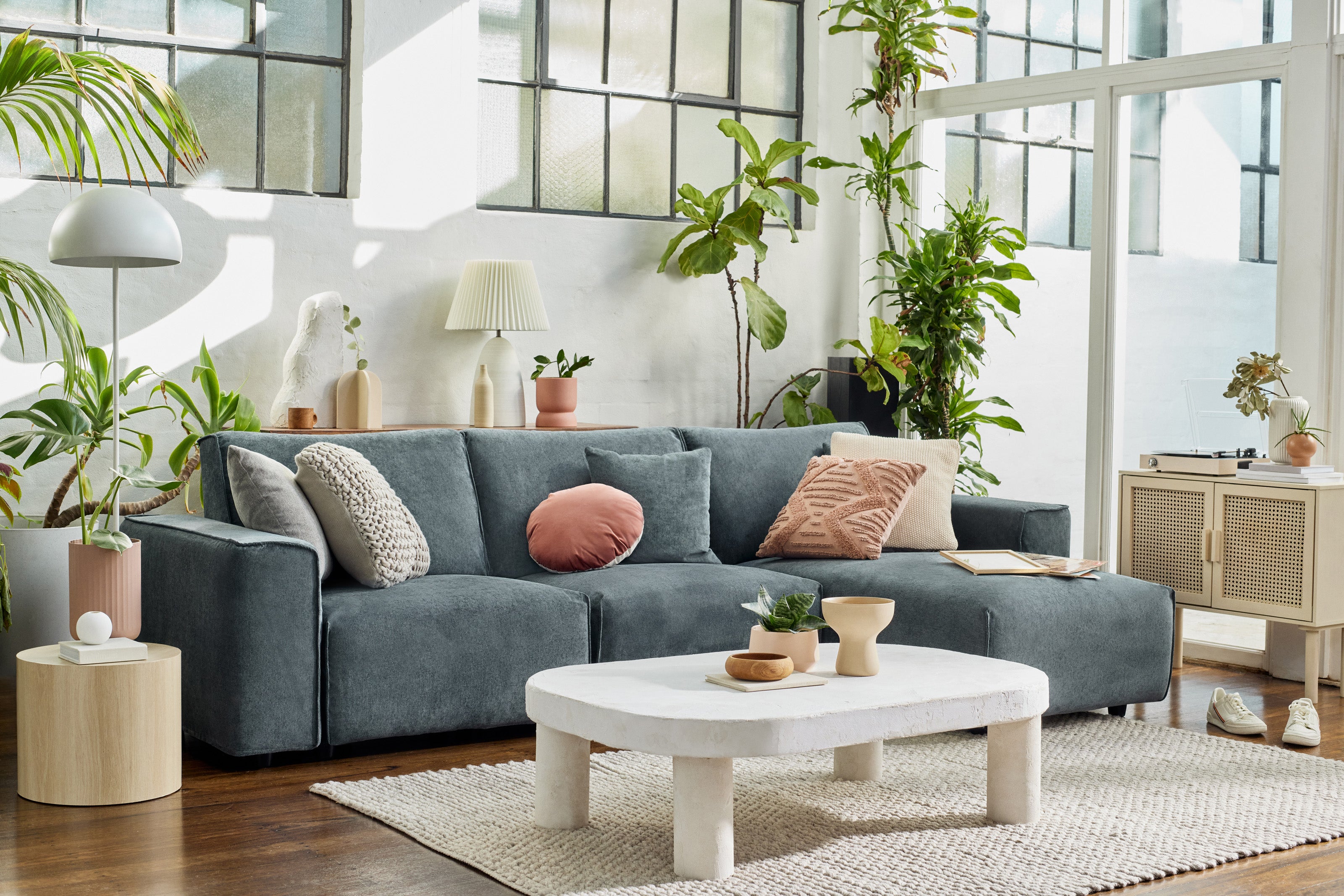 Modern living room with grey sofa, assorted cushions, white coffee table, and potted plants. Large windows and warm lighting create a cosy ambience.