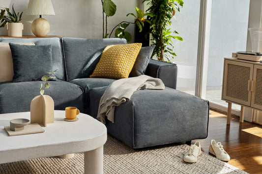 Modern living room with a dark grey sectional sofa, yellow patterned cushion, beige throw, light grey coffee table, and indoor plants by a window.
