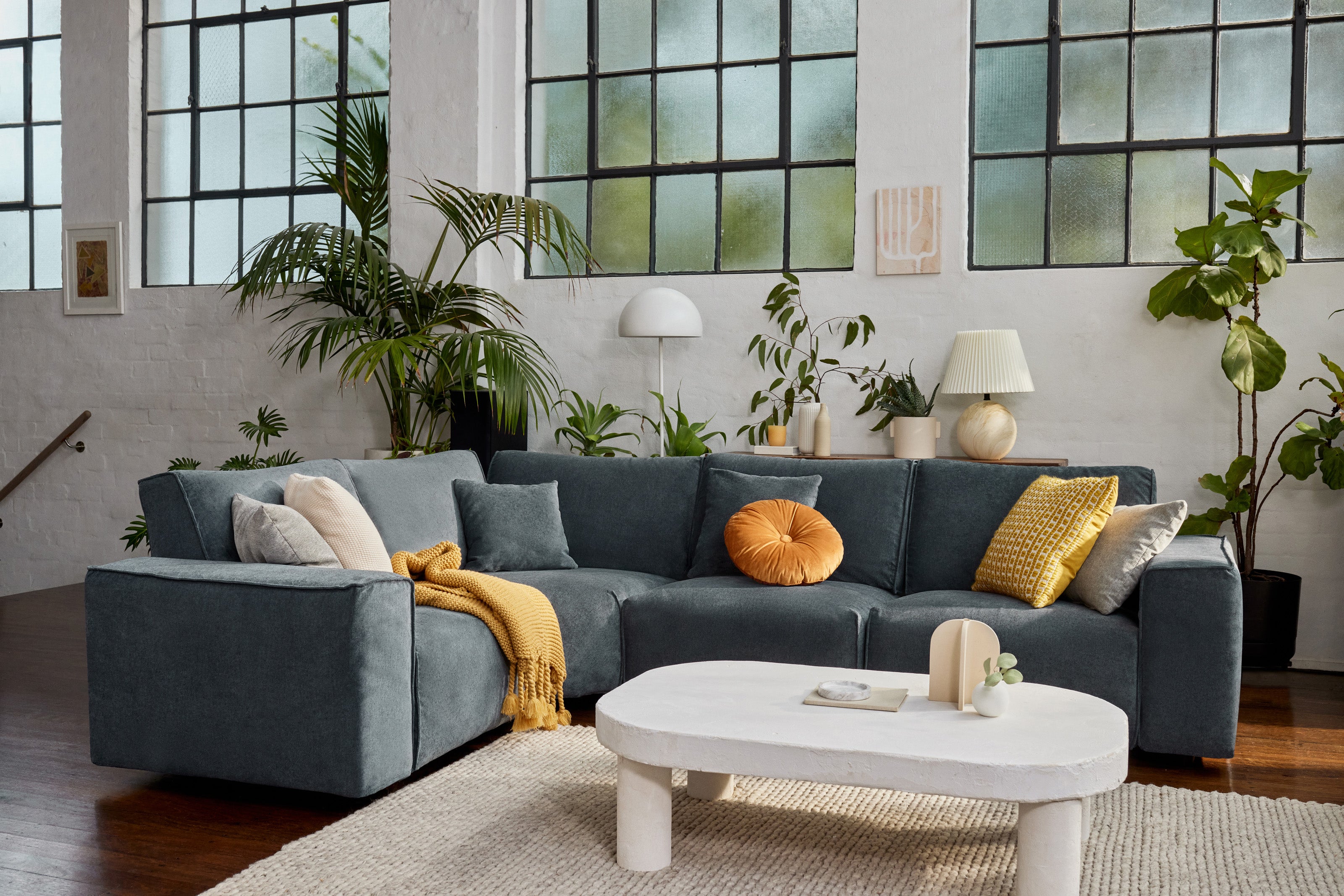 Modern living room with a dark grey L-shaped sofa adorned with orange and yellow cushions, beside a white oval coffee table; lush green plants in the background.