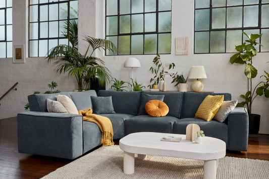 Modern living room with a dark grey L-shaped sofa adorned with orange and yellow cushions, beside a white oval coffee table; lush green plants in the background.