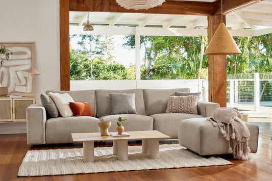 Modern living room with a light grey sectional sofa, neutral cushions, a wooden coffee table, and a woven rug on wooden flooring. Large windows.