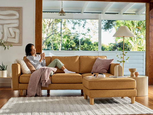 Woman relaxing on mustard-coloured sofa with matching ottoman in bright room. Decor includes a pink throw, soft cushions, and a table lamp.