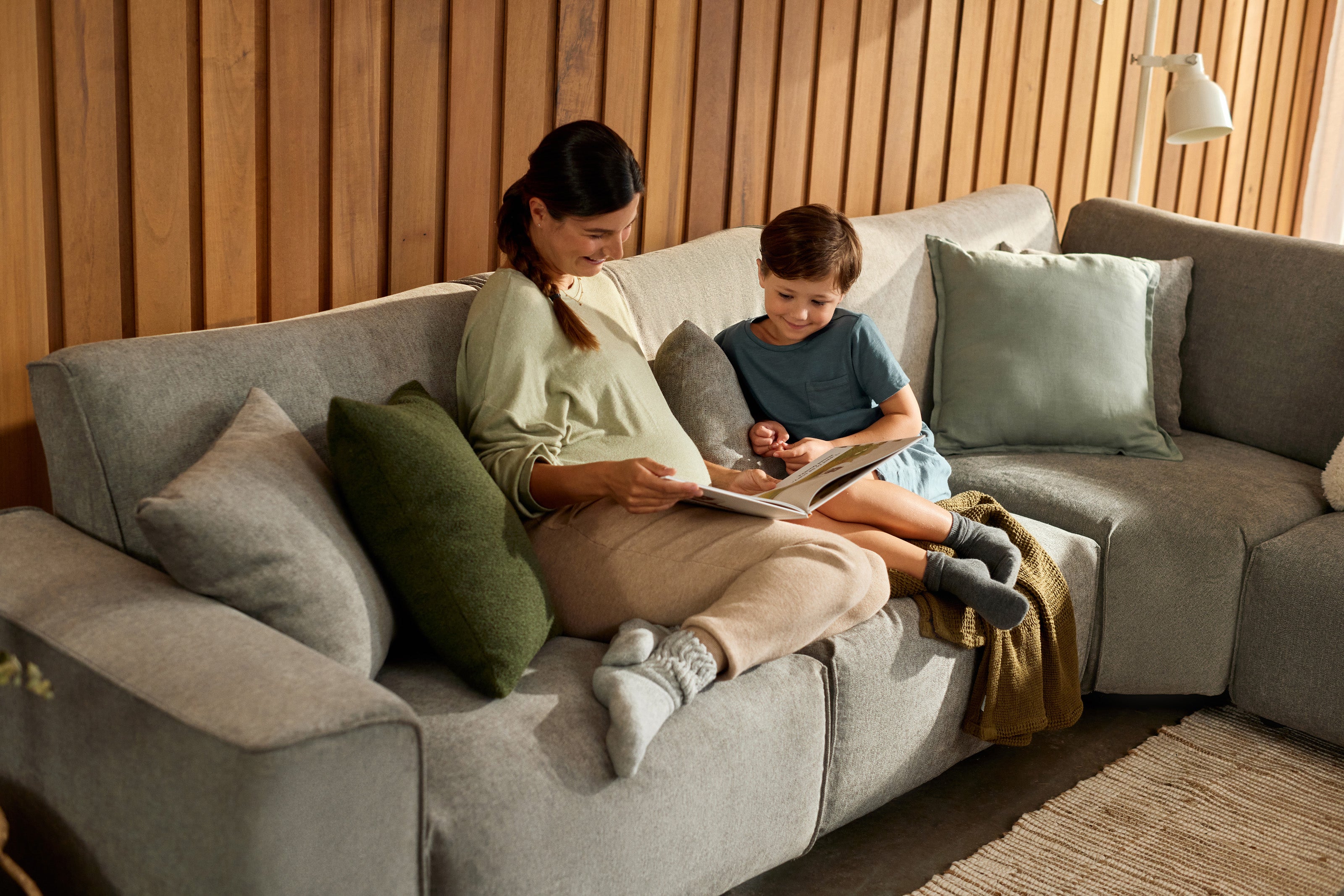 A woman and child sitting on a grey sectional sofa with green and grey cushions, reading a book in a cosy room with wooden wall panelling.