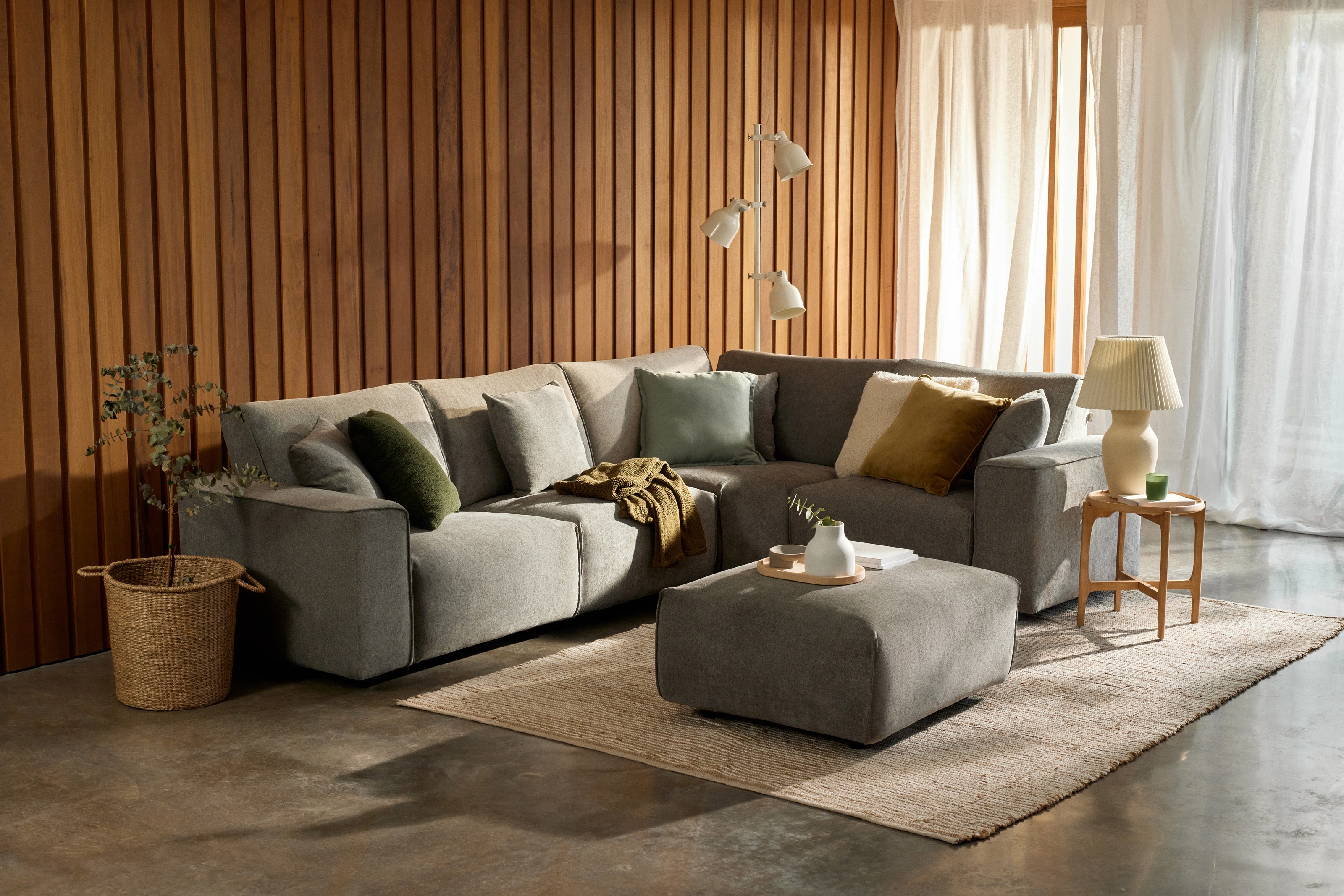 Corner grey sofa with assorted cushions, beige ottoman, and wooden side table in a room with wood-panel walls and sheer curtains.