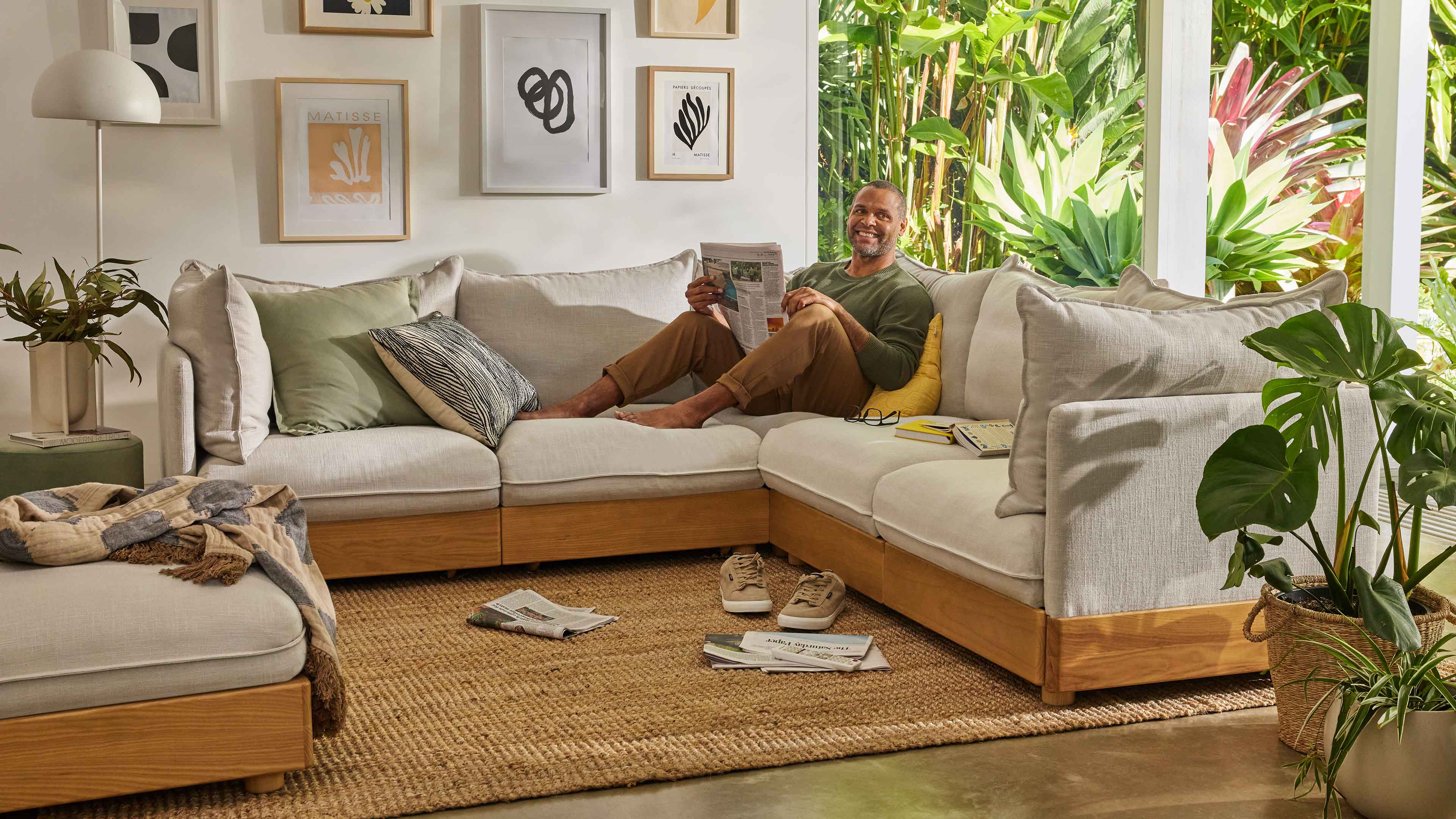 Spacious living room with a light grey sectional sofa on a woven rug, surrounded by plants and artwork. Person reading, shoes and magazines nearby.