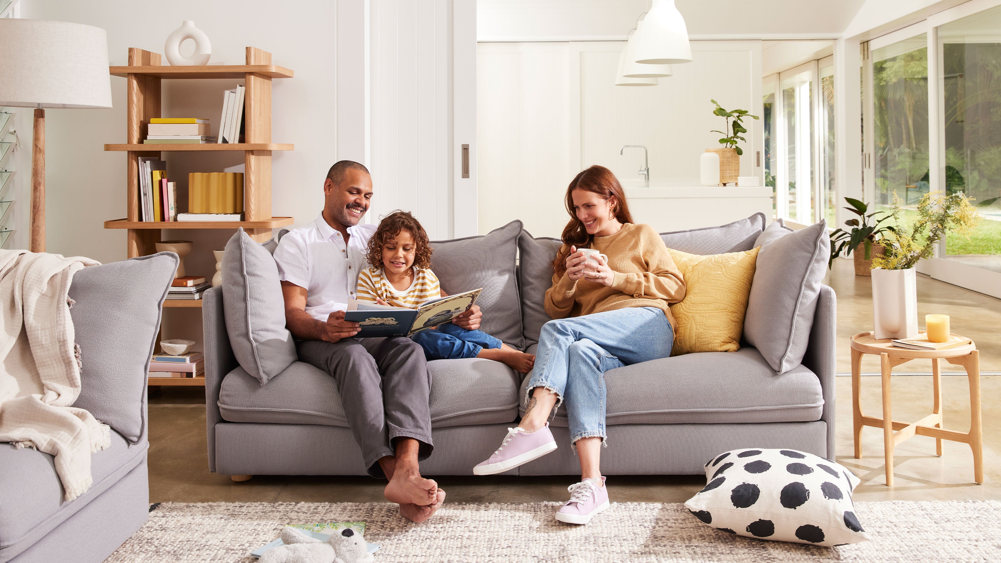 Family relaxing on a grey couch in a bright living room; wooden shelves, yellow cushion, polka dot pillow, natural light, modern decor.