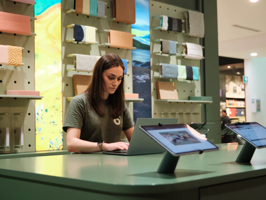 Woman at a green desk using a laptop, with fabric samples and tablets displayed in a showroom; modern interior design setting.