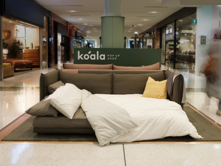 Brown modular sofa in a shopping mall, styled as a bed with white and yellow cushions. "Koala Pop Up Shop" sign in the background.