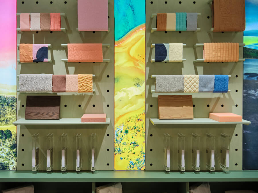 Display of various fabric and wood samples in soft pink, orange, beige, and blue on a pegboard. Background features vibrant abstract artwork.