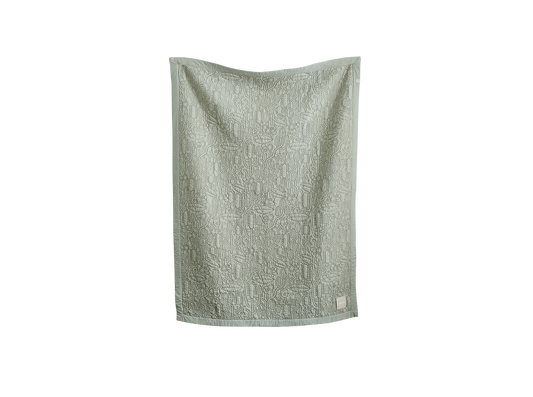 Light green textured quilt hanging, displaying intricate patterns. Fabric label on the bottom right corner.