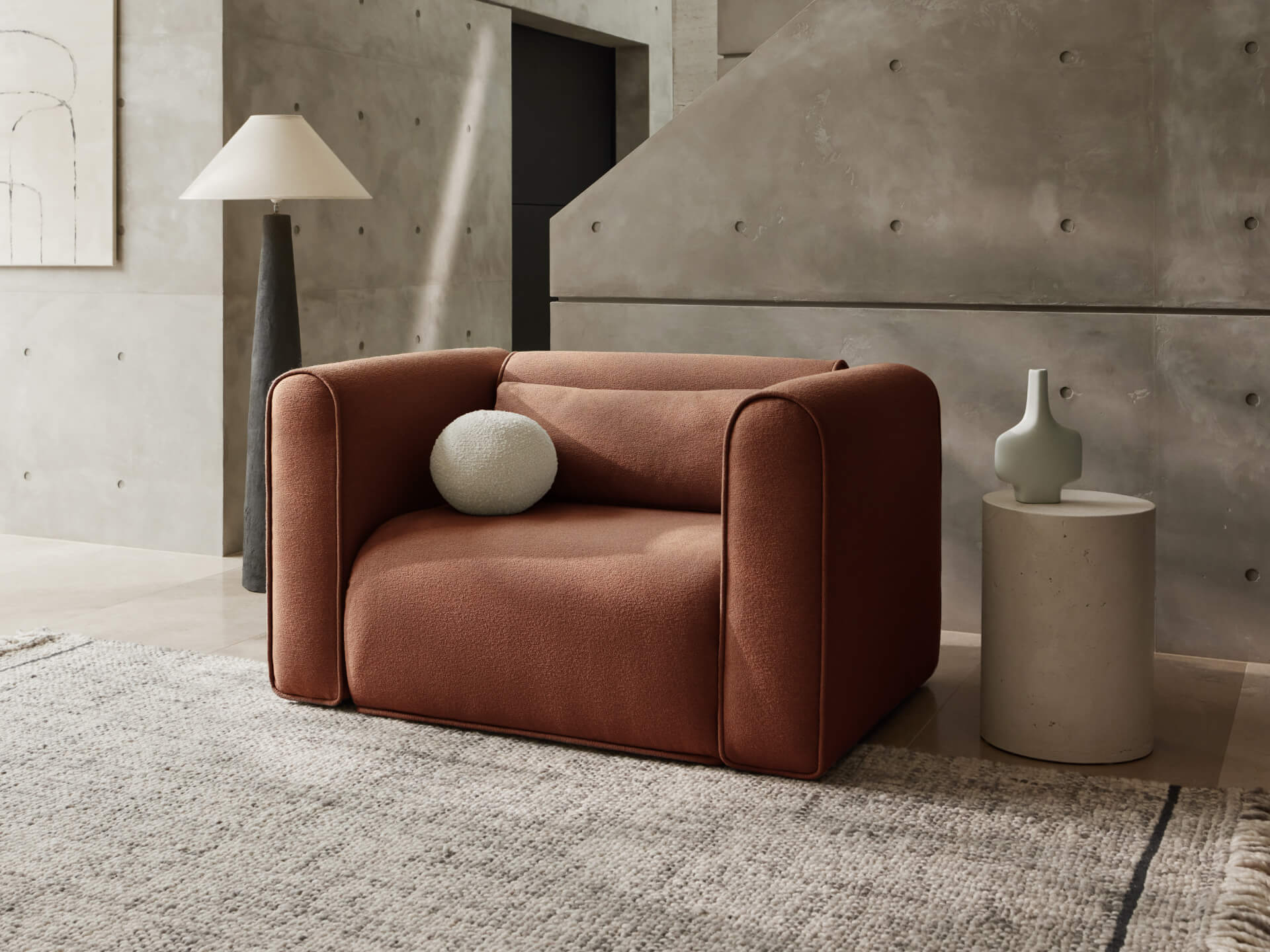 Brown armchair with a round cushion beside a small round table and a grey vase. A textured carpet and tall lamp complete the modern interior.