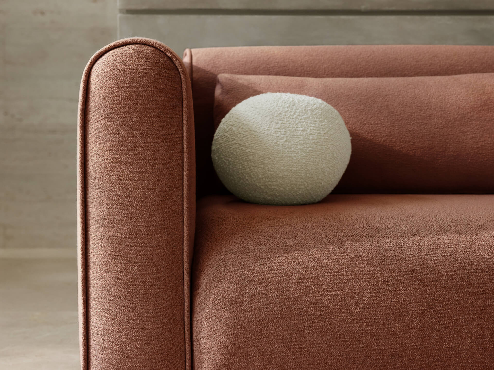 Rust-coloured sofa with rounded armrest and a textured white round cushion, set against a neutral background.
