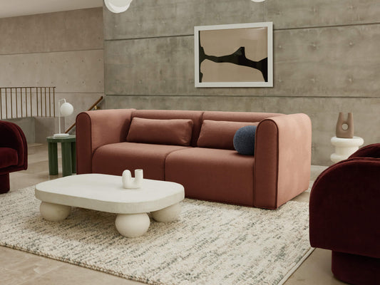 Modern living room with a dusty pink sofa, abstract wall art, cream coffee table on a textured rug, and burgundy chairs. Concrete walls and floor.