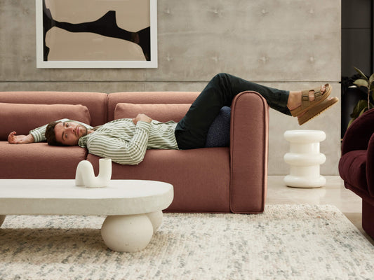 Man relaxes on a pink sofa with feet on the armrest. White coffee table and rug in foreground, abstract wall art behind. Modern decor.