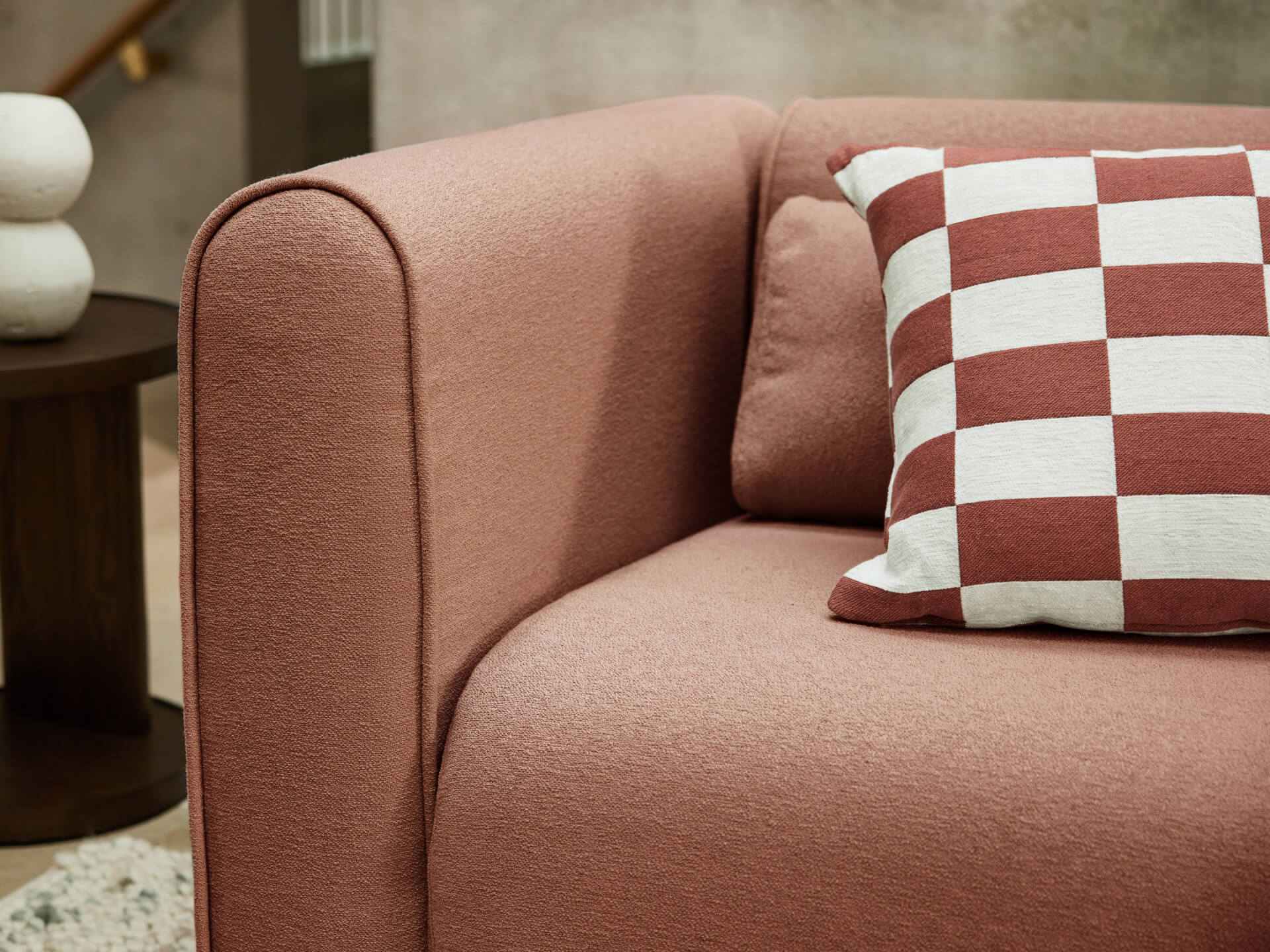 Rust-coloured sofa corner with a white and rust checkered cushion. Dark wooden side table and abstract decor in the background.