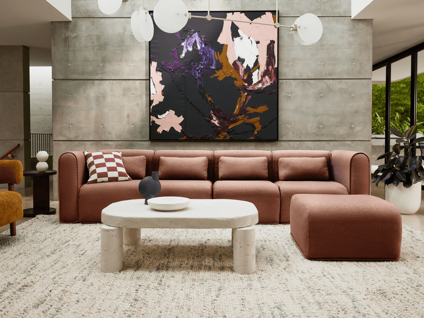 Modern living room with a brown sofa, white oval coffee table, abstract wall art, and a textured rug. Beige walls and green plants in the background.