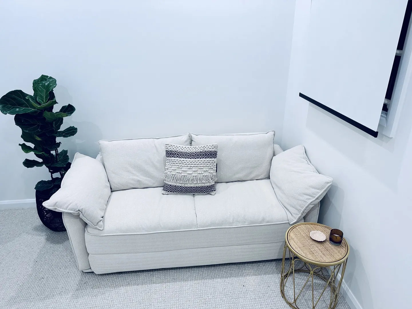 White sofa with textured cushion, small round wicker table, potted plant, and hanging artwork on light carpet in cosy living room corner.