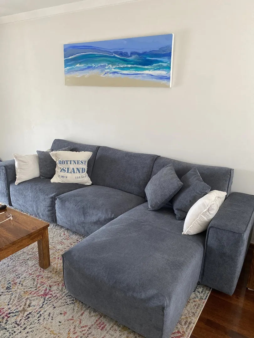 Grey sectional sofa with assorted white and dark grey cushions, set on a patterned rug. A coastal artwork hangs above on a white wall.