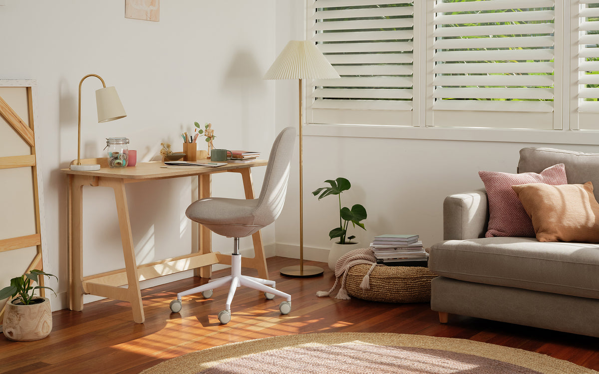 Cosy room with a grey couch, light wood desk, swivel chair, floor lamp, and potted plants. White shutters and soft cushions add warmth.