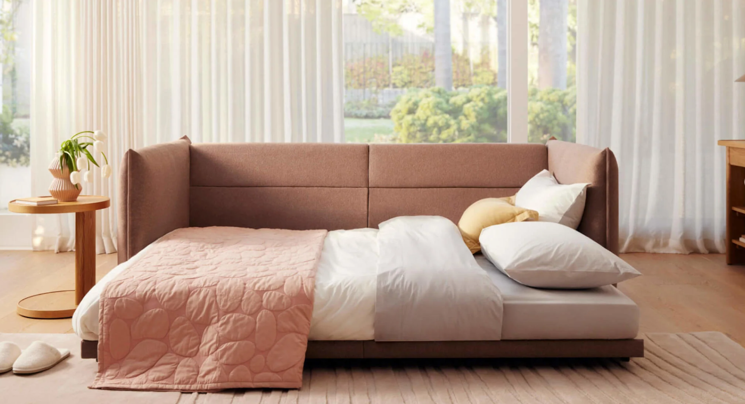 Elegant taupe sofa bed with plush upholstery, set in a sunlit room with floor-to-ceiling windows, complemented by a stylish wooden side table.